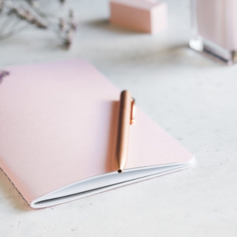 light pink journal with a gold pen resting on a white desk top
