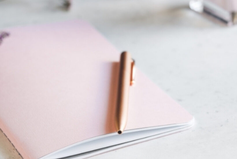 light pink journal with a gold pen resting on a white desk top