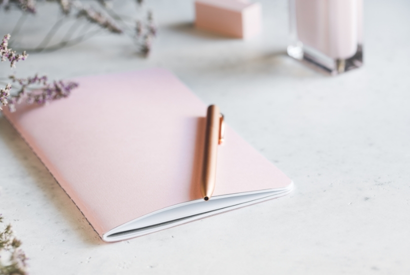 light pink journal with a gold pen resting on a white desk top